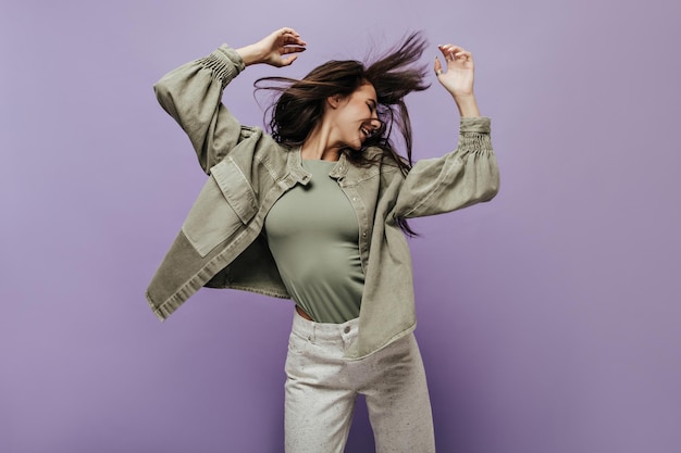 Cheerful girl in fashionable olive top and jacket and cool beige trousers smiling and playing her long dark hair on lilac backdrop