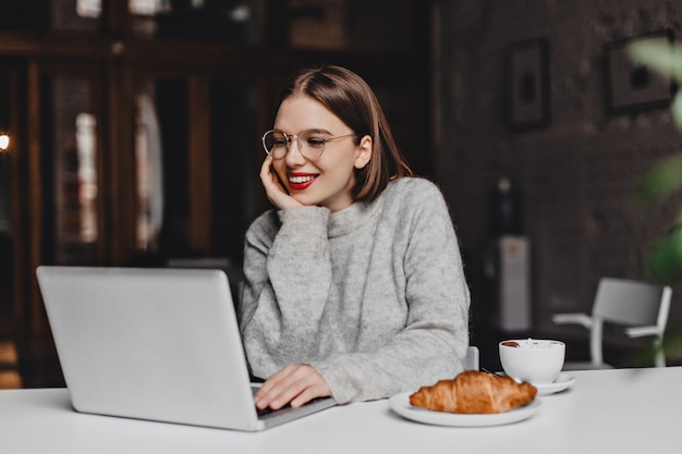 Cheerful girl in gray outfit working in laptop during lunch with croissant and cup of coffee.