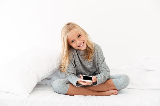 Cheerful girl in gray pajamas holding smartphone while sitting on bed