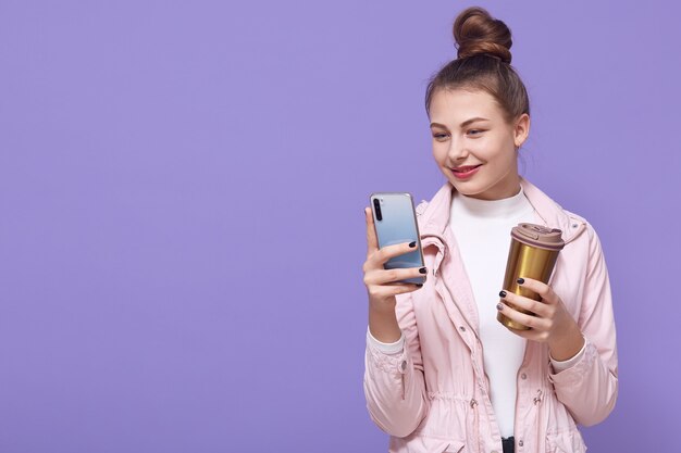 Cheerful girl has coffee break, standing with gadget in hands, reads notification on mobile phone, updates favorite app, types message and smiles while looking at screen, wears jacket, copy space.