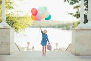 Foto gratuita ragazza allegra che tiene i palloni variopinti e la valigia puerile
