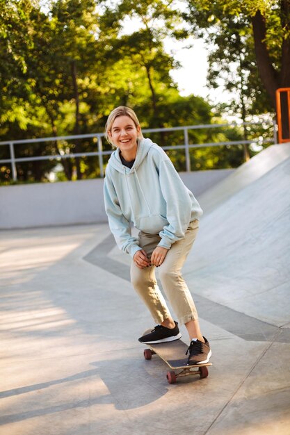 Cheerful girl in hoodie happily looking in camera while trying skateboarding at modern skatepark