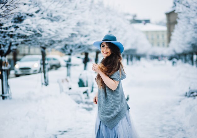 Cheerful girl in light summer dress, sweater and hat walks along the park alley 