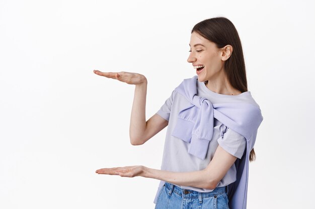 Cheerful girl looking at empty space, holding object box large size, laughing as staring at copyspace, standing over white wall