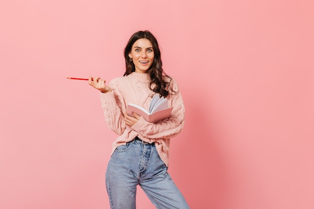 Cheerful girl in mom jeans and pink sweater in amazement posing on isolated background. Woman with wavy dark hairs has thought of what to write in her diary.