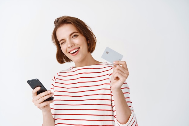 Cheerful girl paying with smartphone online, showing plastic credit card for shopping and smiling, standing against white wall