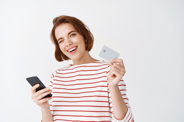 Веселая девушка расплачивается со смартфоном онлайн, показывает пластиковую кредитную карту для покупок и улыбается, стоя у белой стены