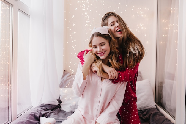 Cheerful girl in pink night-suit sitting on bed with eyes closed. Indoor photo of joyful european woman embracing her sister with happy smile.
