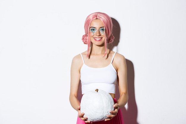 Cheerful girl in pink wig and halloween costume holding white pumpkin, smiling and celebrating holiday