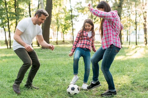 陽気な少女が公園で芝生の上の彼女の両親とサッカーボールをプレー