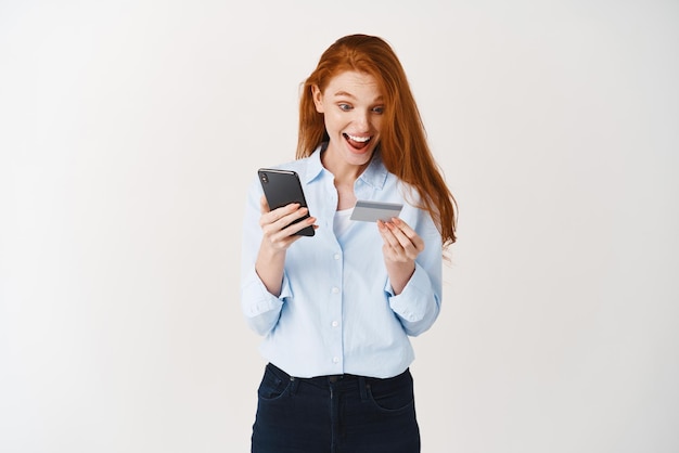 Cheerful girl shopping online making purchase in smartphone app with plastic credit card standing against white background