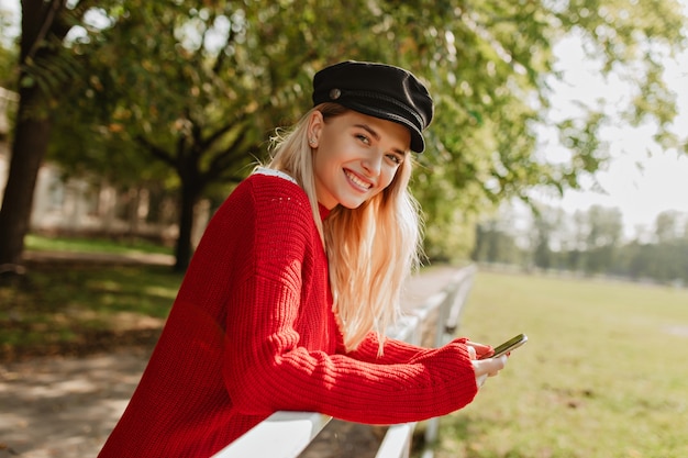 Веселая девушка, красиво улыбаясь, хорошо проводит время в парке. Привлекательная блондинка в красивой красной сезонной одежде.