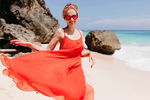 Cheerful girl in sparkle sunglasses posing with sincere smile near rocks on sea. Photo of pleasant young woman expressing happiness in sunny day at ocean beach.