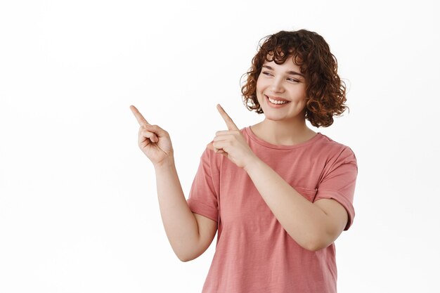 Cheerful girl student with curly hairstyle, pointing and looking at upper left corner with pleased white smile, happy gazing at copy space, standing on white