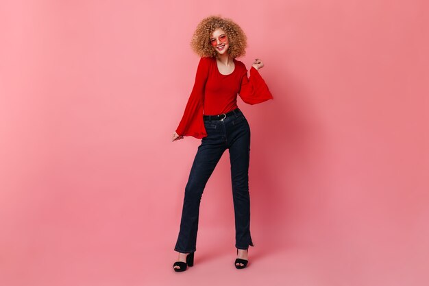 Cheerful girl in stylish red top and dark denim pants dancing. Portrait of curly lady in glasses on pink space.