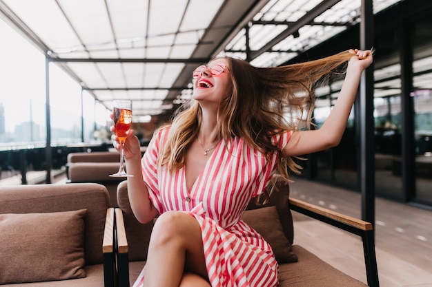 Cheerful girl in stylish striped dress having fun in cafe and drinking cocktail. Laughing blonde caucasian woman playing with her hair while posing in restaurant.