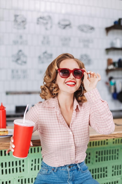 Cheerful girl in sunglasses and shirt sitting at the bar counter with soda water in hand and happily spending time in cafe