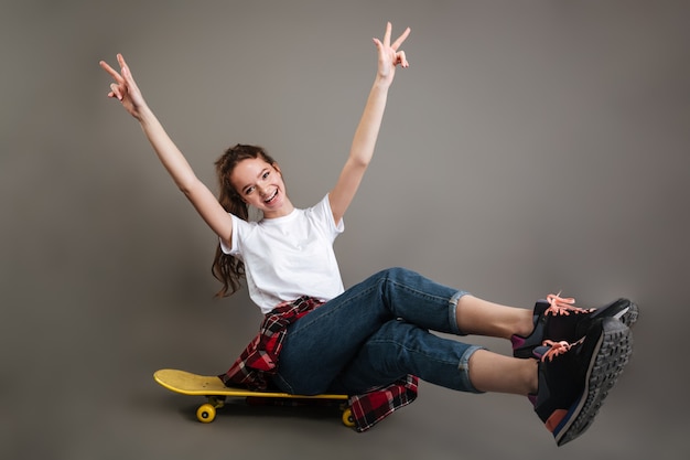 Cheerful girl teenager sitting on skateboard and showing peace sign