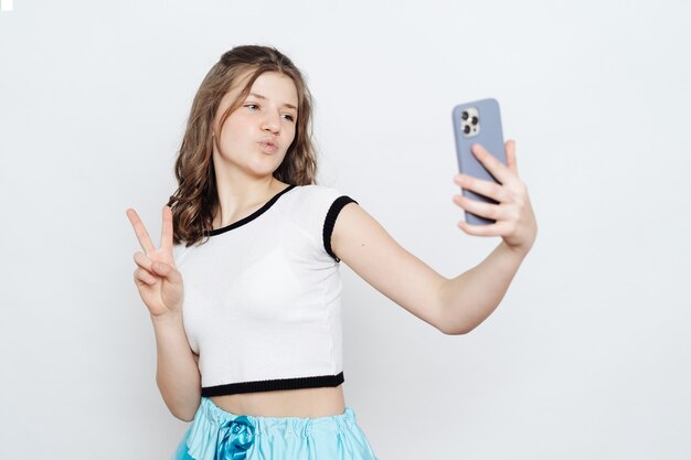 Веселая девочка-подросток, делающая селфи с телефоном, позирует на белом
