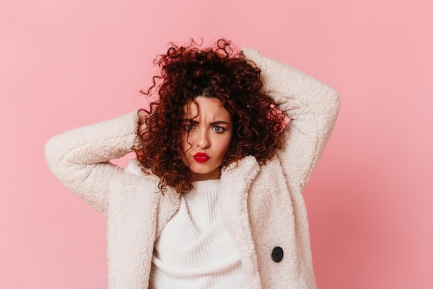 Cheerful girl in white warm outfit tousles her curly dark hair and looks into camera on pink background