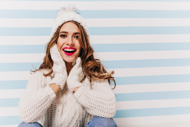 Ragazza allegra con grandi occhi belli in posa con eccitazione in divertente cappello lavorato a maglia. ritratto dell'interno del magnifico modello femminile indossa guanti e maglione ridendo