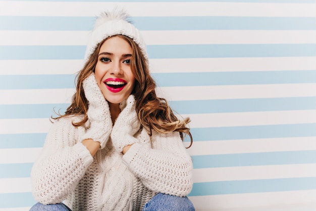 Cheerful girl with big beautiful eyes posing with excitement in funny knitted hat. Indoor portrait of magnificent female model wears gloves and sweater laughing