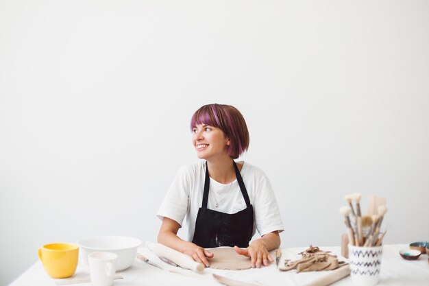 Cheerful girl with colorful hair in black apron and white T-shirt working with clay happily looking aside at modern pottery studio