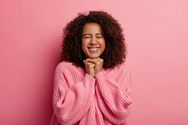 cheerful girl with crisp hair, keeps hands together under chin, happy to achieve goal, has eyes closed, smiles broadly isolated on pink studio wall