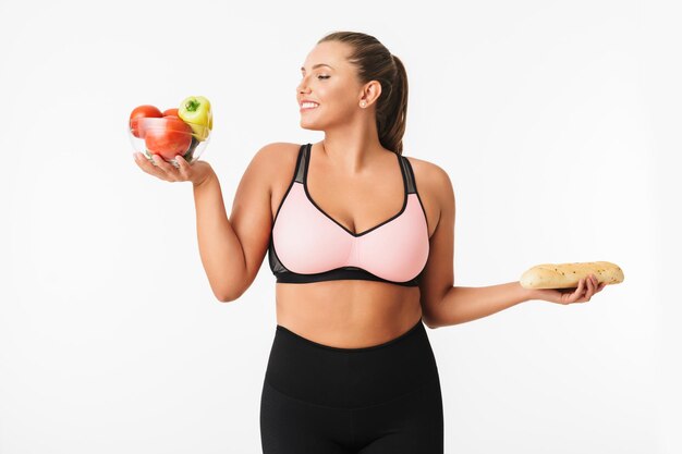 Cheerful girl with excess weight in sporty top happily holding bowl with vegetables and bread in hands over white background isolated