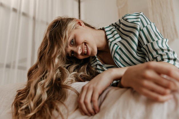 Cheerful girl with long curly hair looks down into camera smiles and resting on big comfortable so