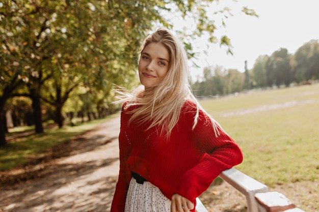 Жизнерадостная девушка с естественным макияжем радостно улыбается. Довольно блондинка счастливо позирует в осеннем парке.