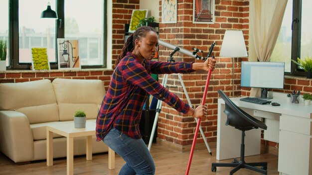 Cheerful girlfriend dancing in apartment and using mop to sweep floors, listening to music and having fun spring cleaning. Positive young woman singing and mopping tiles at home.