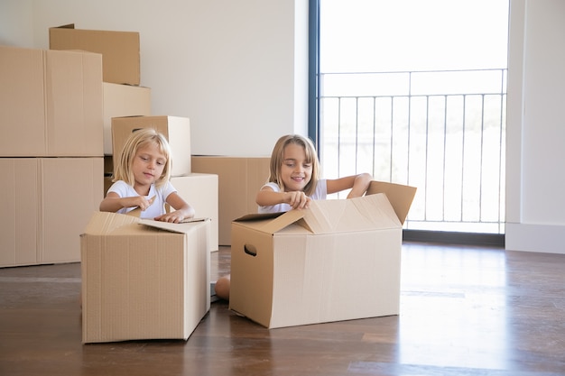 Cheerful girls unpacking things in new apartment, sitting on floor and opening cartoon boxes