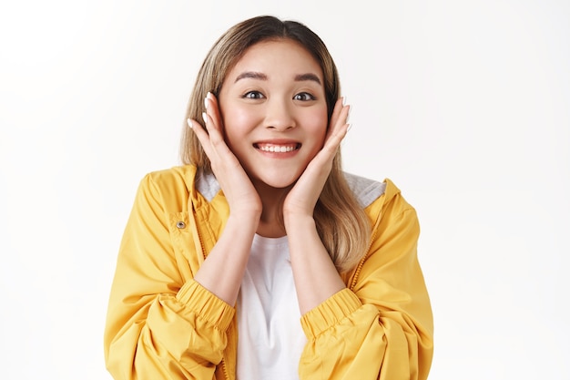 Cheerful glad pretty shiny smiling happy asian blond girl touch cheeks surprised receive positive great news expressed excitement delighted grin thrilled thankful triumphing, white wall