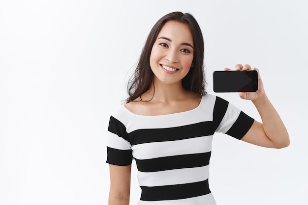 Allegra felice giovane donna asiatica in maglietta a righe che tiene lo smartphone orizzontalmente vicino alla testa inclinata del viso e sorridente amichevole consiglia di utilizzare l'app che mostra l'applicazione in piedi sullo sfondo bianco