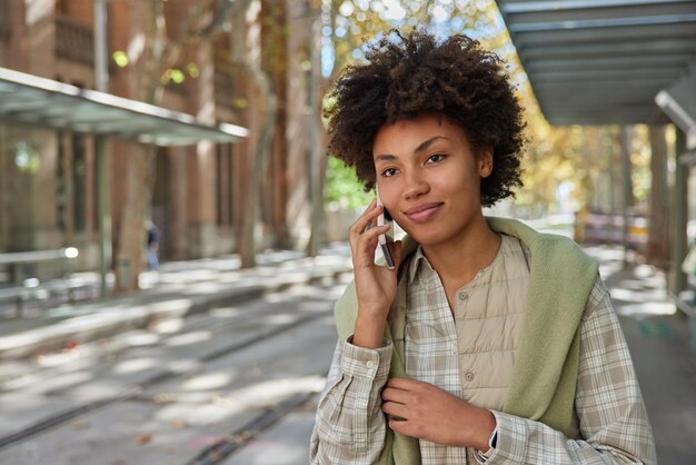 陽気な格好良い縮れ毛の女性は、友人がスマートフォンを使用しているのを聞いて幸せです国際的なオペレーターの関税を使用していますカジュアルな服を着ています