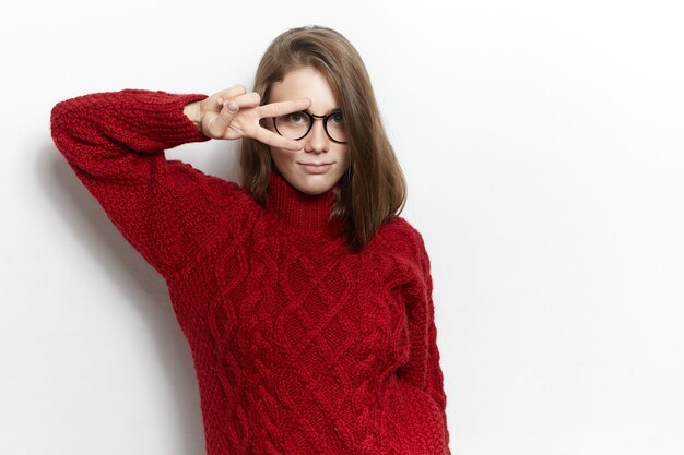 Cheerful good looking cute young woman in spectacles and knitted turtleneck sweater, making V-sign with two fingers over one eye gesture