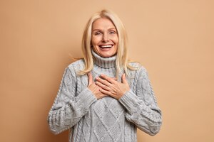 Cheerful good looking middle aged woman keeps hands pressed on chest smiles broadly and expresses positive emotions dressed in winter jumper happy to hear compliment.