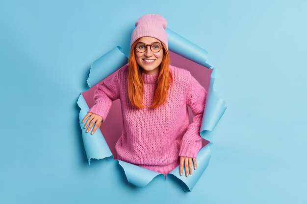 Cheerful good looking woman smiles broadly has red hair dressed in casual clothes has happy mood hears excellent news breaks through blue paper. Joyful ginger millennial girl indoor
