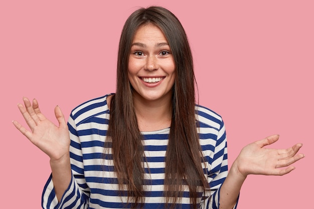 Cheerful good looking young Caucasian woman with long dark straight hair, feels puzzled what she wants