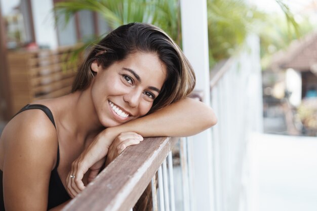 Cheerful good-looking young tanned woman lean on terrace rail and gazing happily