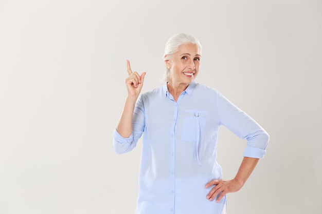 Cheerful gray-haired old lady in blue shirt, pointing with finger upward