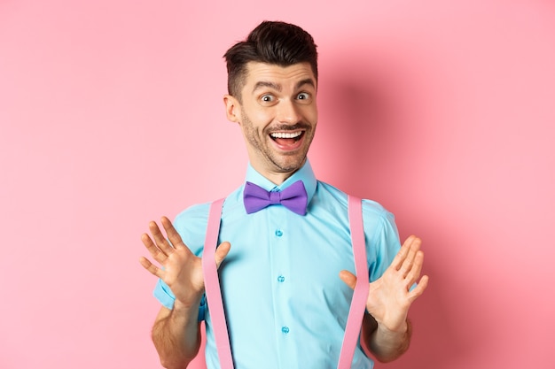 Веселый парень в галстуке-бабочке, показывая свои подтяжки и улыбаясь счастливым, чувствуя оптимизм, стоя возбужденным на розовом фоне.
