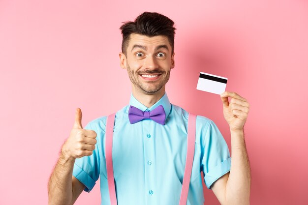 親指を立ててプラスチックのクレジットカードを見せて、プロモオファーのように、カメラで幸せそうに笑って、ピンクの背景の上に立っている蝶ネクタイの陽気な男。