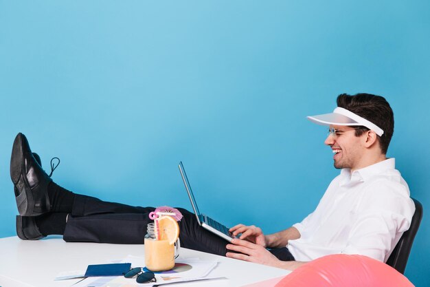 Веселый парень в кепке работает за ноутбуком смеется сидит с ногами на столе Мужчина в офисной одежде работает в отпуске