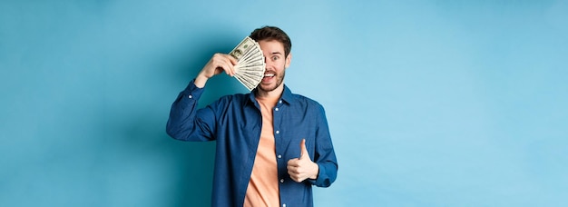 Free photo cheerful guy cover half of face with money and showing thumbsup recommending fast cash loan standing