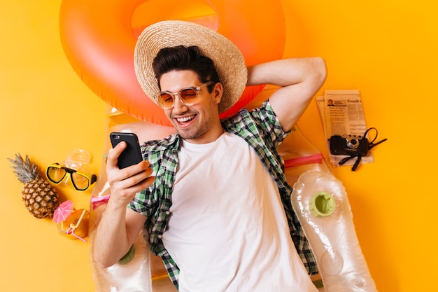 Cheerful guy in hat is chatting on phone while lying on inflatable mattress on orange space.