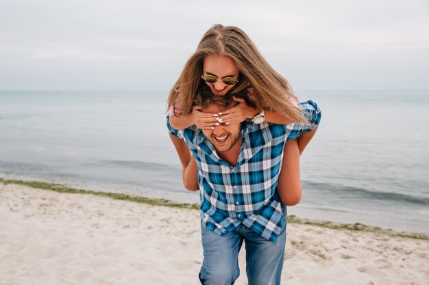 Cheerful guy holding her girlfriend behind the back, while she covering his eyes by hands