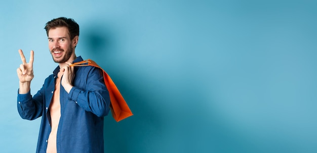 Cheerful guy holding orange shopping bag on shoulder smiling and showing peace sign standing on blue