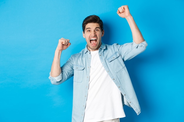 Cheerful guy making fist pumps and rooting for someone, shouting for joy, triumphing over win, standing against blue background